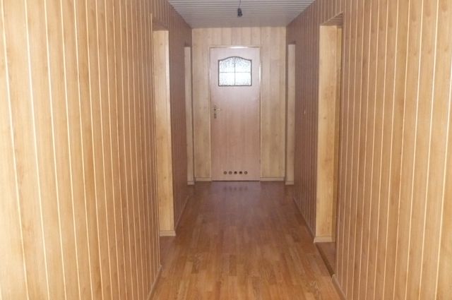 Korytarz wykończony panelami ściennymi w odcieniach jasnego drewna, z drewnianą podłogą i drzwiami z szybą w centralnej części kadru.