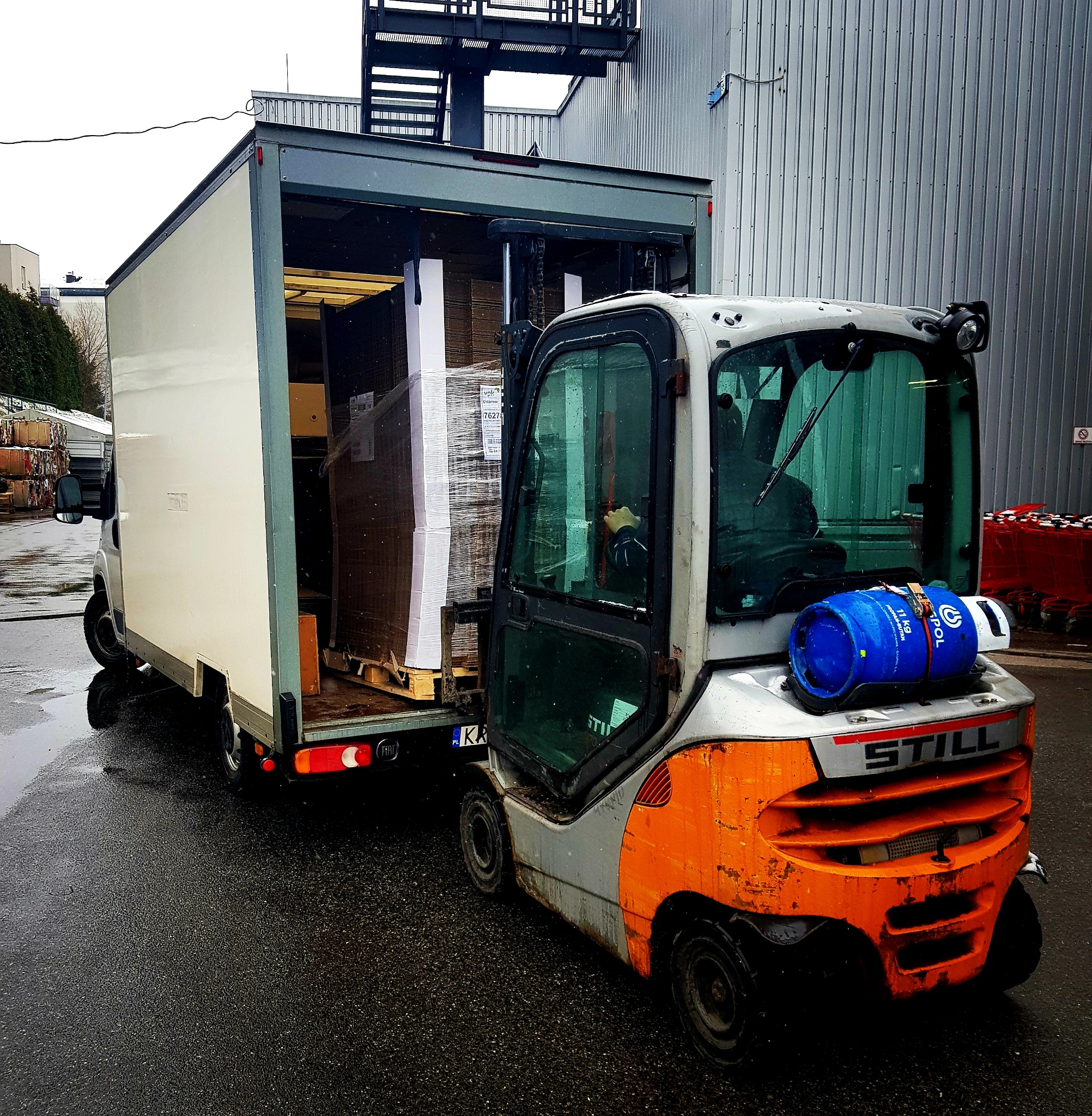 Wózek widłowy STILL ładuje paletę z towarem do białej furgonetki z otwartymi drzwiami na mokrym parkingu przemysłowym.