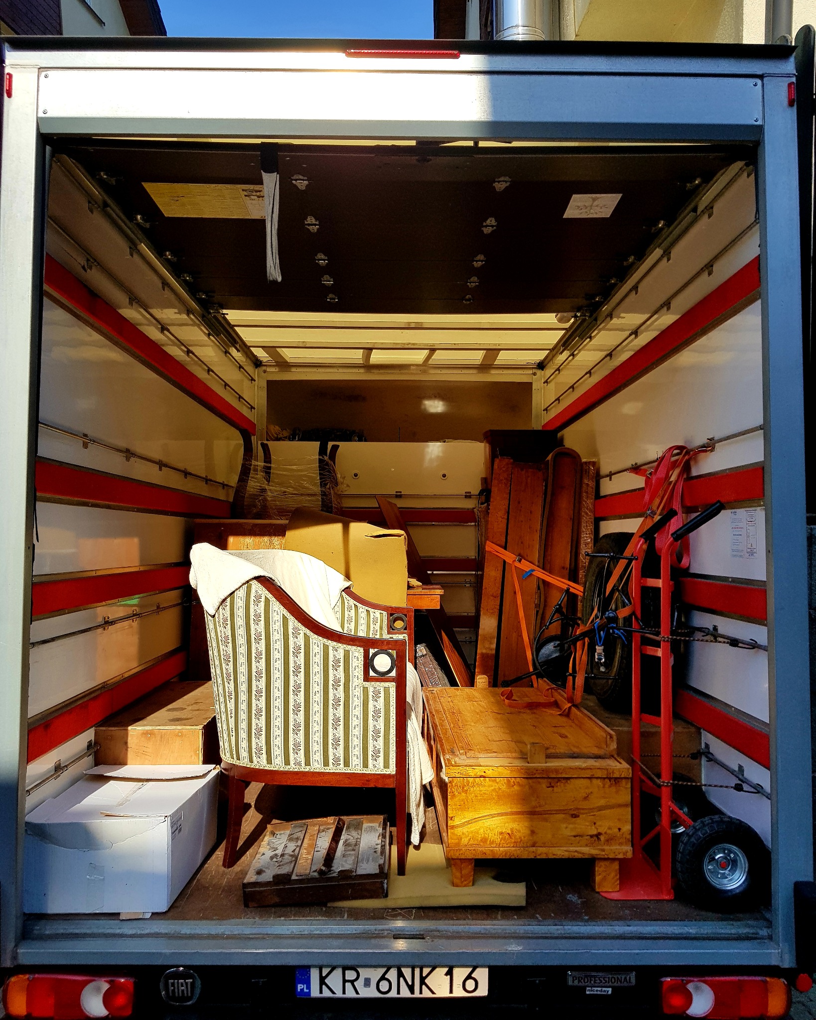Wnętrze częściowo załadowanego busa z meblami antykami, w tym fotel w paski, drewniana skrzynia i wózek transportowy z pasami mocującymi.