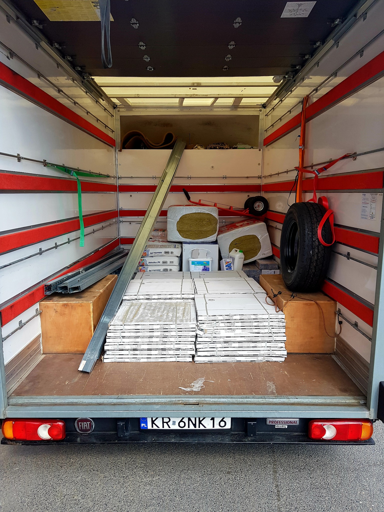 Wnętrze furgonetki załadowane materiałami budowlanymi: płyty gipsowe, wełna mineralna, profile metalowe, zapakowane w kartony i worki, zabezpieczone pasami transportowymi.
