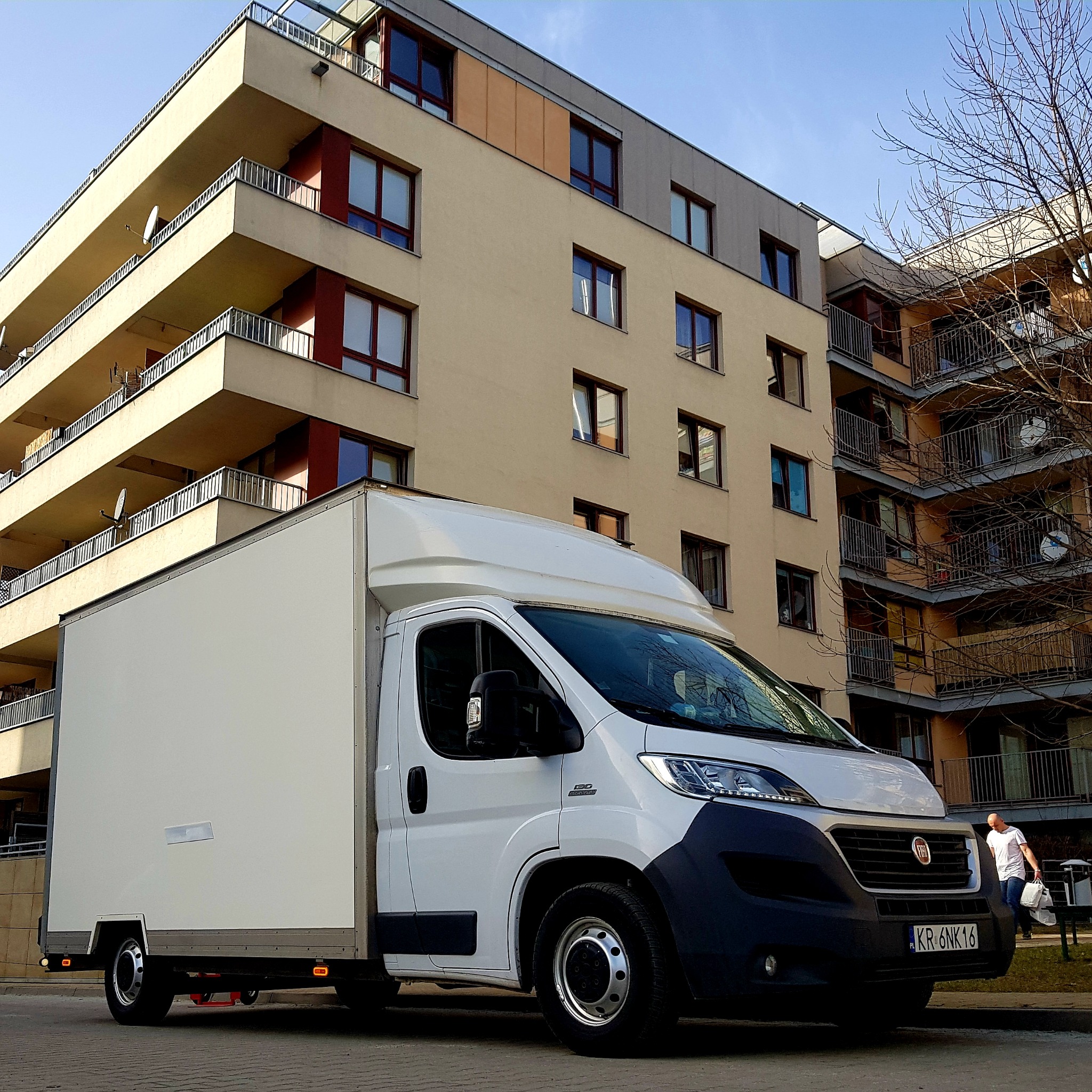 Biały samochód dostawczy Fiat Ducato z zabudową kontenerową na tle nowoczesnego budynku mieszkalnego z balkonami i antenami satelitarnymi.