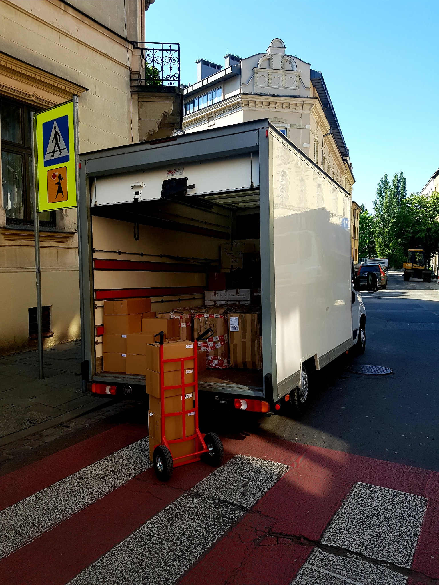 Ciężarówka z otwartą przestrzenią ładunkową wypełnioną kartonami, wózek transportowy z kartonami na pasach dla pieszych, znak drogowy w tle.