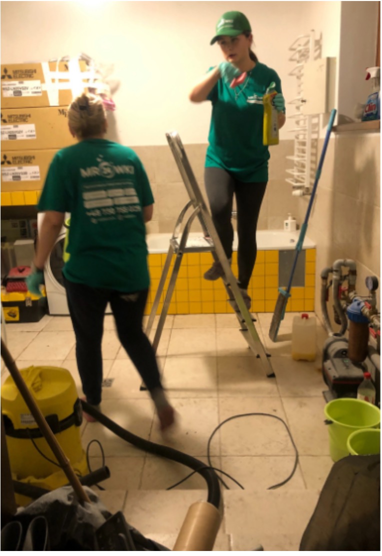 Dwie osoby w zielonych uniformach czyszczą łazienkę z żółtymi płytkami, jedna na drabinie z butelką środka czyszczącego, druga z odkurzaczem przemysłowym, widoczne narzędzia i środki czystości.