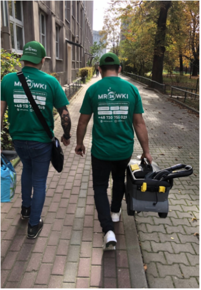 Dwaj mężczyźni w zielonych koszulkach z logo firmy idą chodnikiem, jeden niesie odkurzacz do prania tapicerki.
