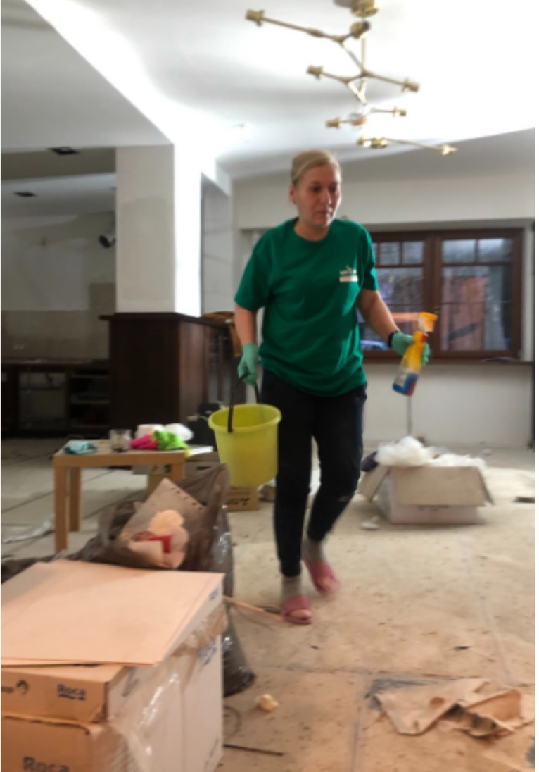 Pracownica w zielonej koszulce z logo, niosąca żółte wiadro i butelkę ze spryskiwaczem, w pomieszczeniu w trakcie sprzątania, widoczne kartony, śmieci i resztki po remoncie.