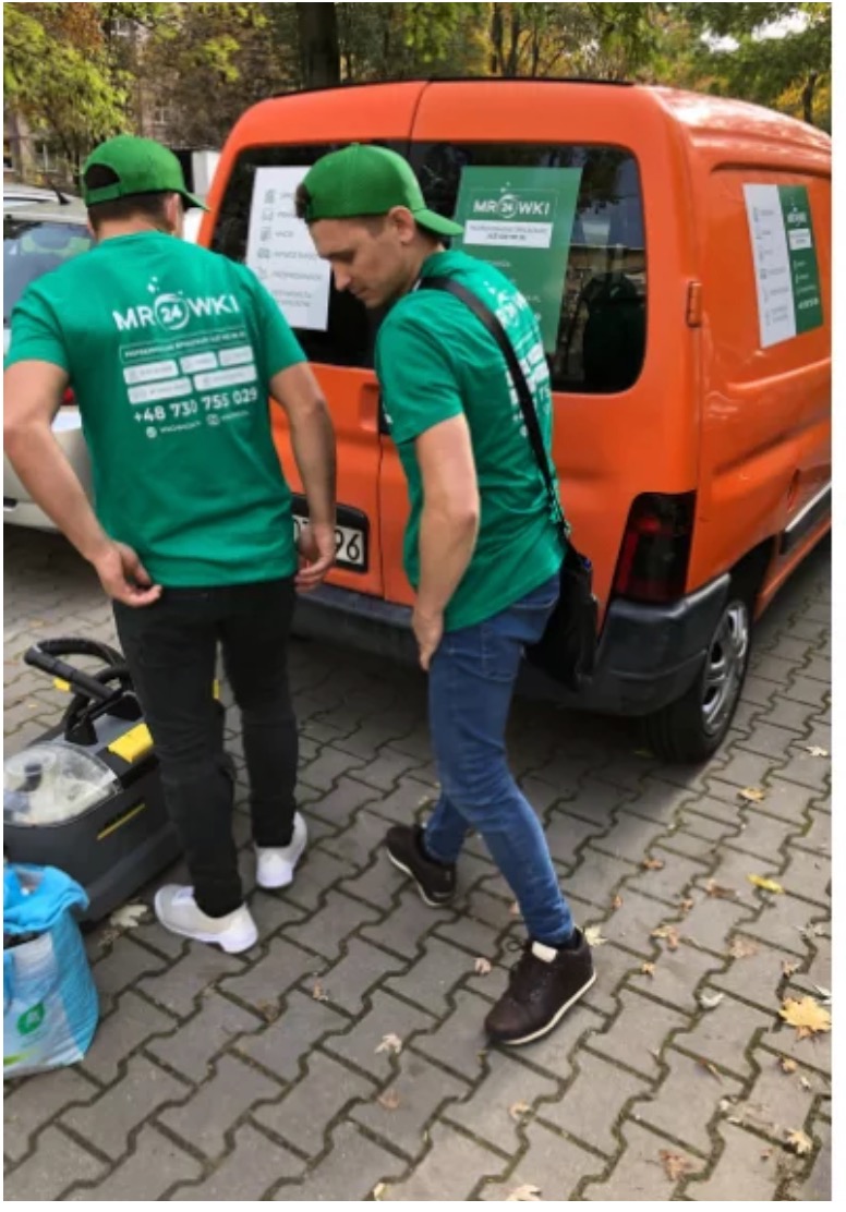 Dwaj mężczyźni w zielonych koszulkach firmowych stojący obok pomarańczowego samochodu dostawczego z reklamą firmy, na chodniku leży profesjonalny odkurzacz piorący.