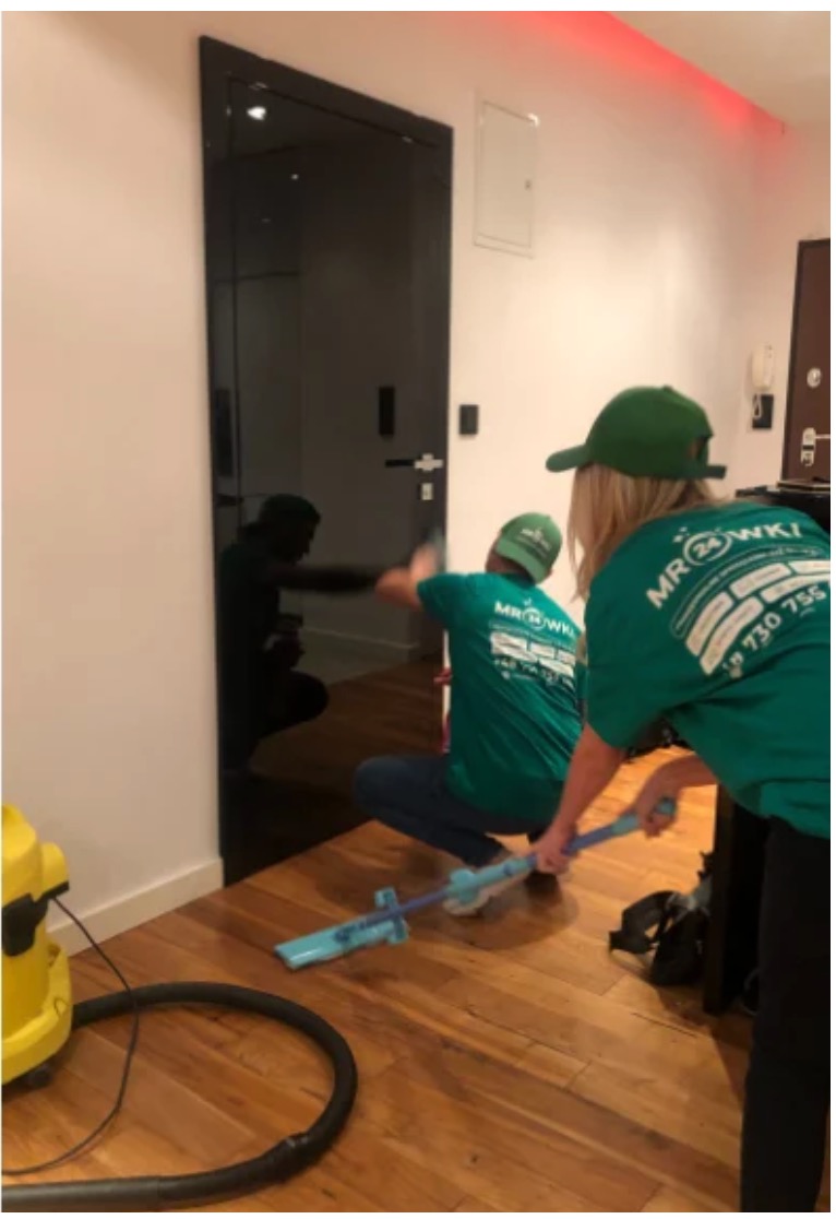 Dwie osoby w zielonych koszulkach firmowych czyszczą czarną, szklaną taflę drzwiową w pomieszczeniu z drewnianą podłogą, używając niebieskiego mopa do podłóg i odkurzacza.