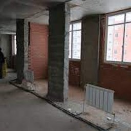 Roman Sokolov - Surowe wnętrze w trakcie remontu biura: widoczne betonowe kolumny, fragmenty ściany z czerwonej cegły, grzejnik, okna z widokiem na budynek, materiały budowlane, instalacja elektryczna na podłodze...