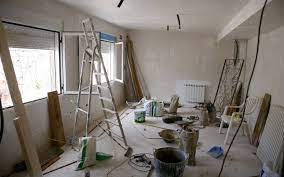 Pokój w trakcie remontu: widoczne drabiny, worki z zaprawą, narzędzia malarskie, niepomalowane ściany i okna z częściowo zasłoniętymi roletami.