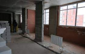 Surowe wnętrze w trakcie remontu biura: widoczne betonowe kolumny, fragmenty ściany z czerwonej cegły, grzejnik, okna z widokiem na budynek, materiały budowlane, instalacja elektryczna na podłodze...