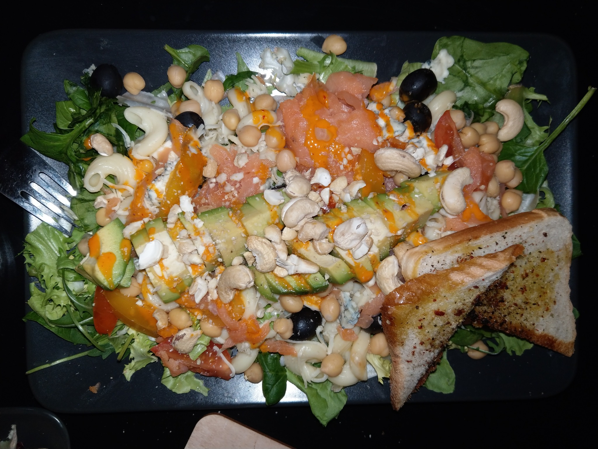 Sałatka z łososiem, awokado, ciecierzycą, makaronem, orzechami nerkowca, oliwkami, pomidorami i grzankami na czarnym talerzu, polana sosem.