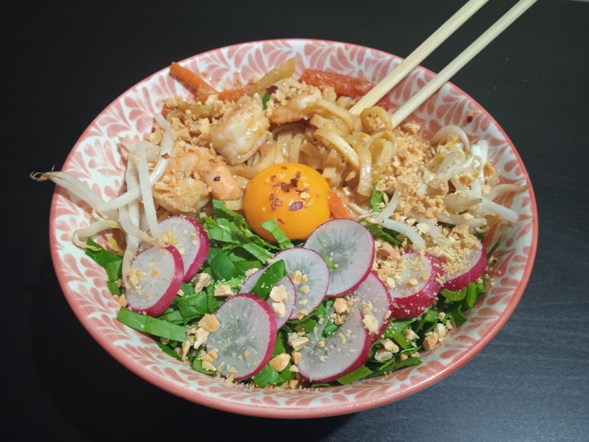 Pad Thai z krewetkami, rzodkiewką, kiełkami fasoli mung, orzeszkami ziemnymi i żółtkiem na wierzchu, podane w różowej misce z pałeczkami.