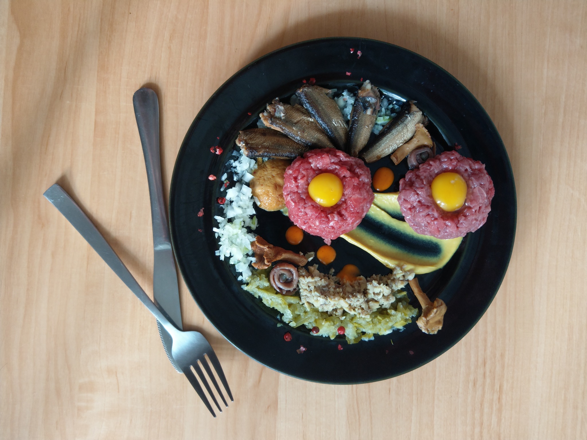 Czarny talerz z tatarem wołowym z żółtkiem, śledziami, siekaną cebulą, sosem, grzybami i ogórkami na drewnianym stole, obok widelec i nóż.