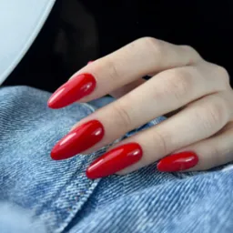 Dłoń z długimi, czerwonymi paznokciami w kształcie migdałów, połyskującymi w świetle lampy UV, na tle dżinsowej tkaniny.