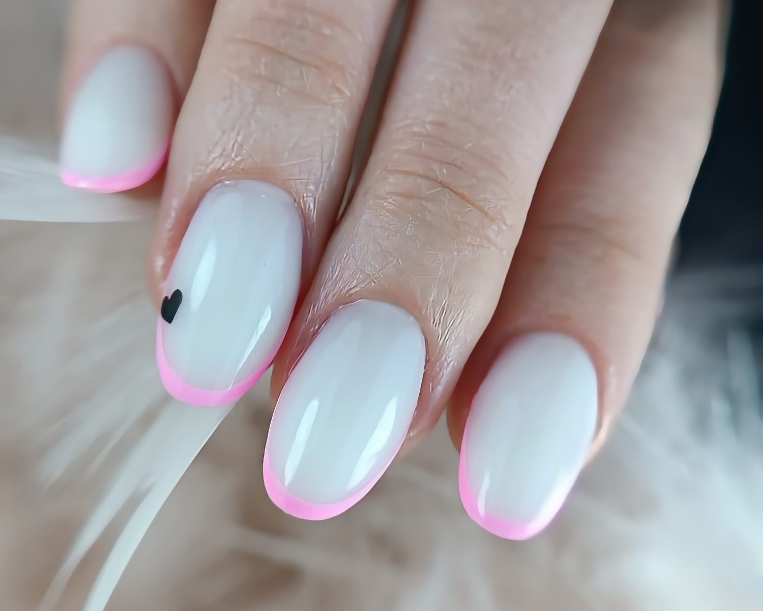 Delikatny manicure: mleczna baza, różowy french i małe czarne serduszko na jednym paznokciu, uchwycone z bliska na tle białych piór.