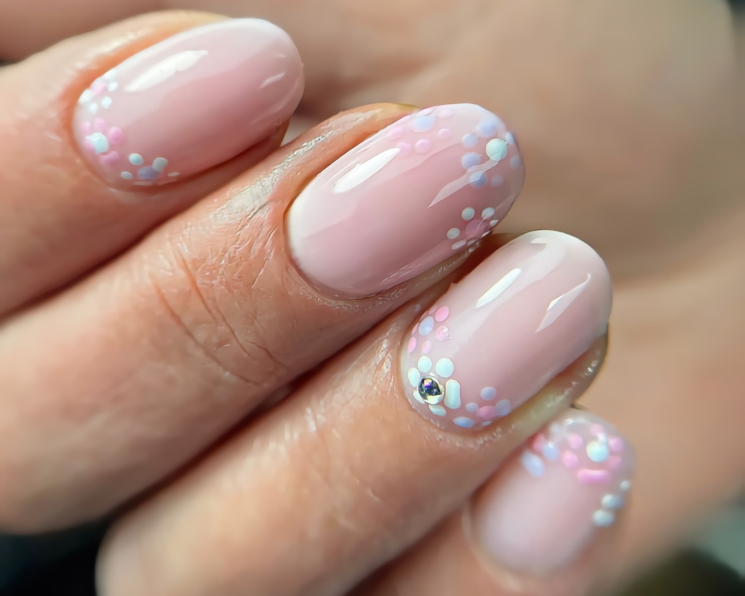 Delikatny, pastelowy manicure żelowy z motywem drobnych, kolorowych kropek przypominających kwiaty; jeden paznokieć ozdobiony małym, błyszczącym kamieniem.