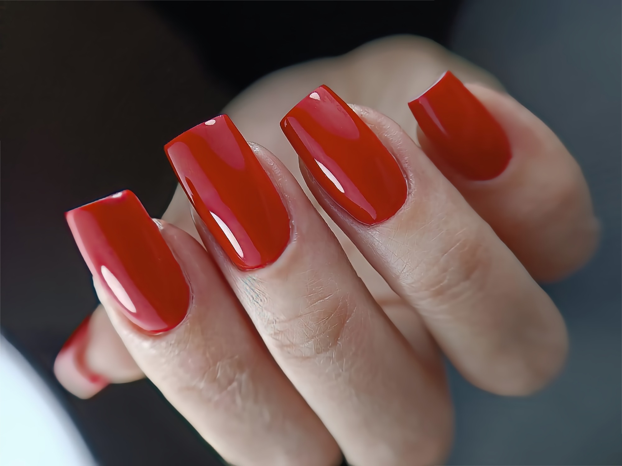 Dłoń z pomalowanymi na intensywną czerwień, kwadratowymi paznokciami o błyszczącym wykończeniu, widoczne delikatne linie papilarne na skórze dłoni.