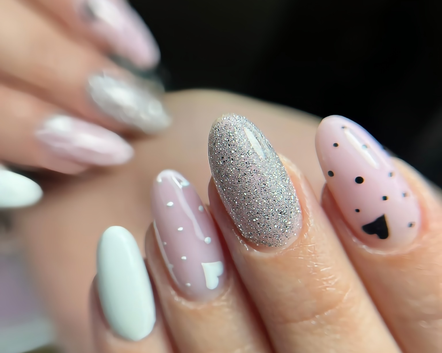 Delikatny manicure walentynkowy: różowe paznokcie z czarnymi serduszkami i białymi kropkami, jeden paznokieć pokryty srebrnym brokatem, zaokrąglony kształt.