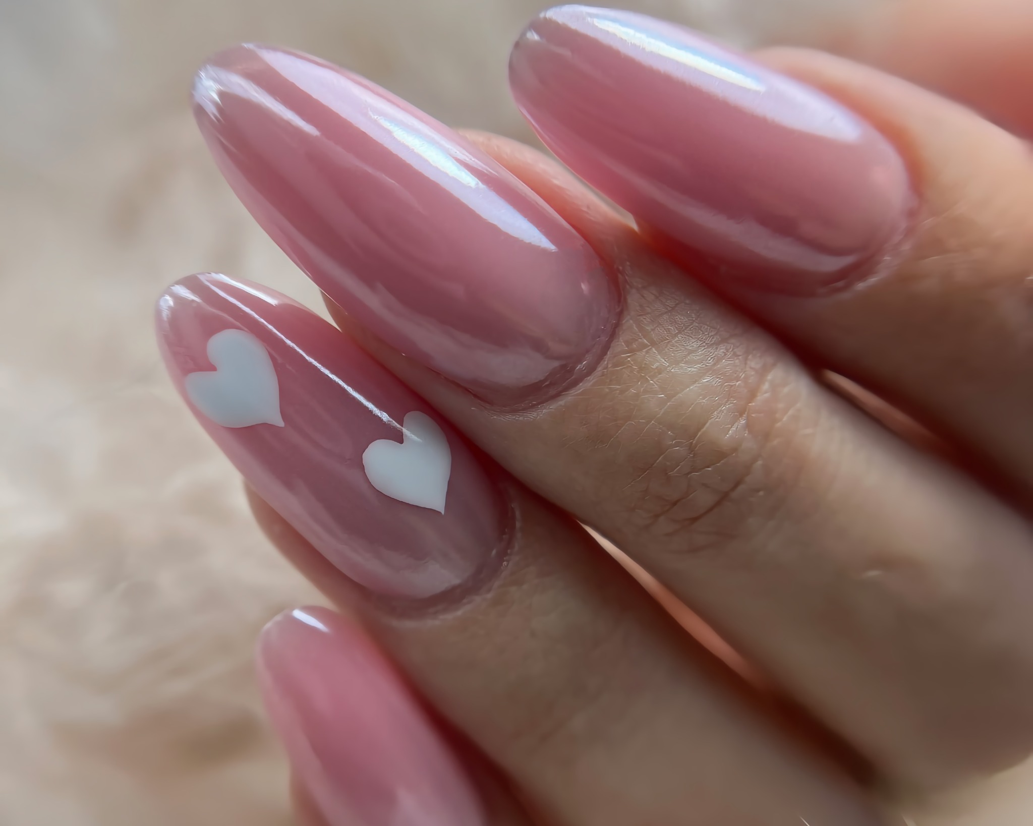 Delikatne, różowe paznokcie w kształcie migdałów z dwoma białymi serduszkami na jednym paznokciu, prezentowane na tle miękkiej tkaniny.