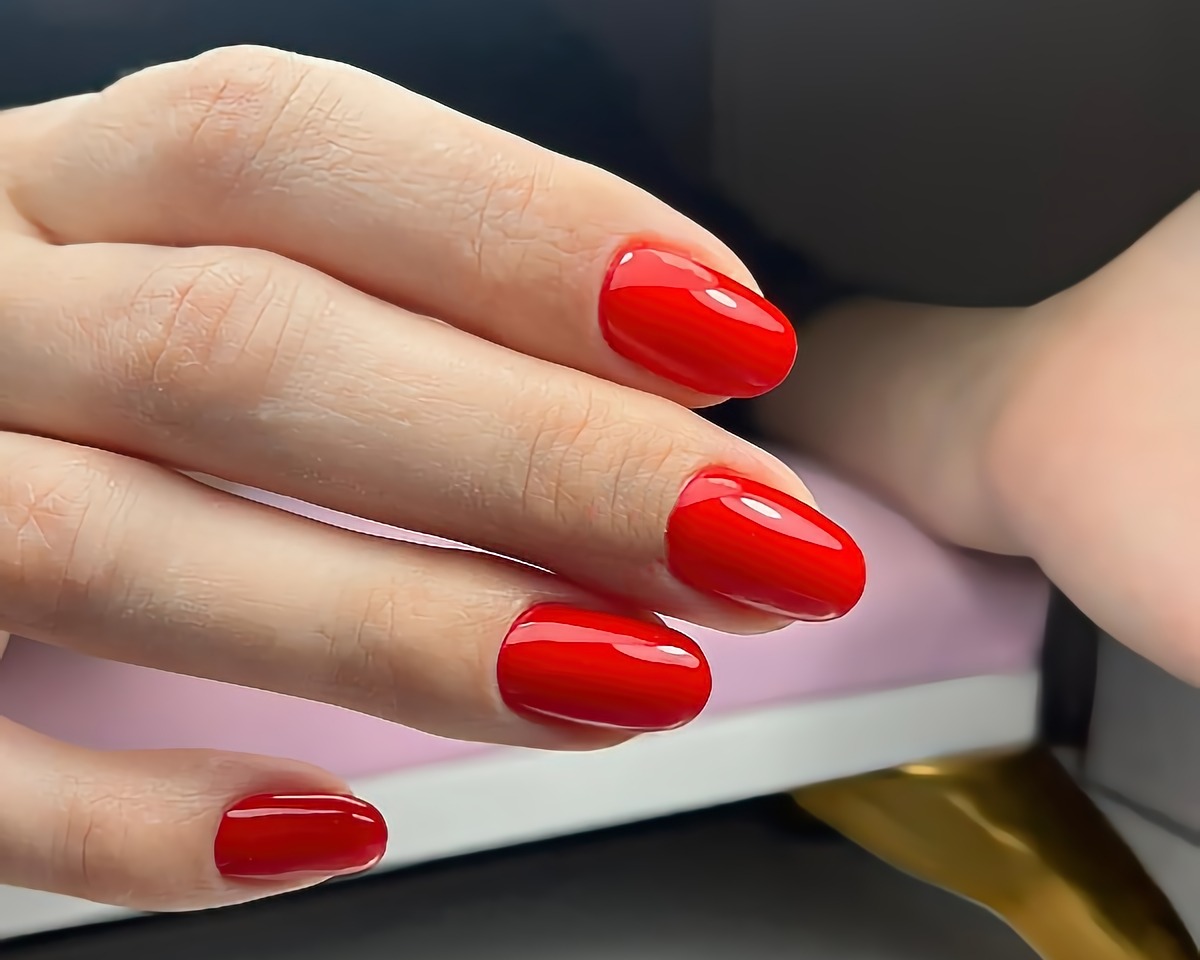 Dłoń z krótkimi, zaokrąglonymi paznokciami pomalowanymi intensywnym czerwonym lakierem hybrydowym, widoczne delikatne odbicie światła na powierzchni paznokci.