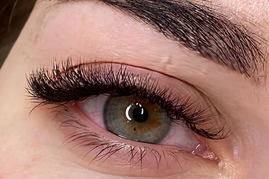 Zbliżenie na oko z wyraźnie zagęszczonymi, przedłużonymi rzęsami, widoczne pojedyncze rzęsy dolne, tęczówka zielono-brązowa z ciemną plamką.