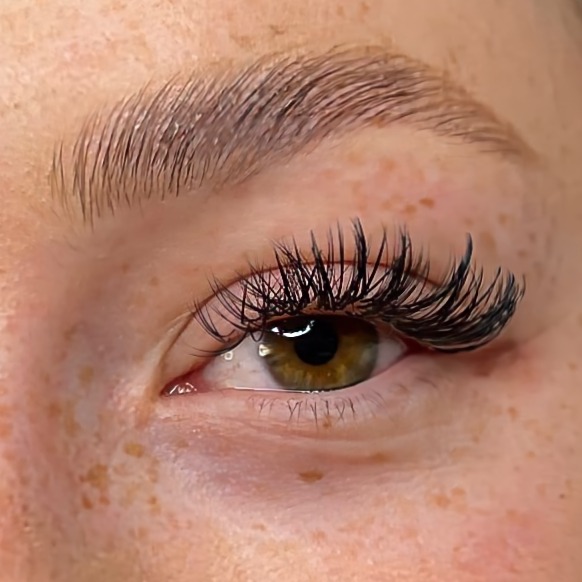 Zbliżenie na oko z przedłużonymi, gęstymi rzęsami w stylu 'mokrego spojrzenia', widoczne piegi na skórze pod okiem i na powiece, brwi naturalne, nieumalowane.