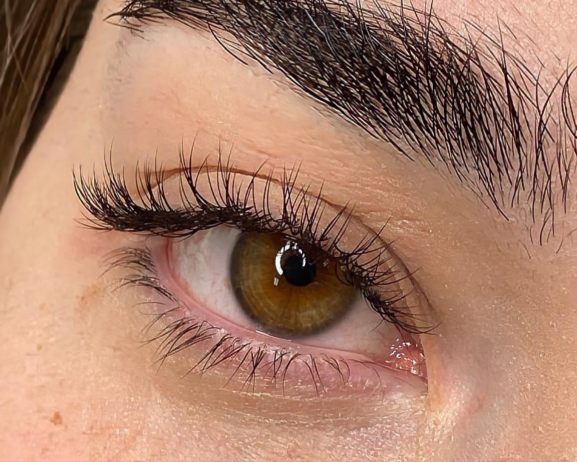 Zbliżenie na oko z przedłużonymi, gęstymi, czarnymi rzęsami. Widoczna brązowa tęczówka i ciemna brew. Skóra wokół oka o jasnym kolorycie z delikatnymi piegami.