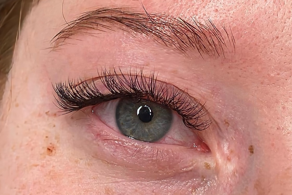 Zbliżenie na oko z wyraźnie przedłużonymi, ciemnymi rzęsami metodą 2:1, widoczne delikatne piegi na skórze wokół oka i brwi.