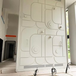 MD Construction - Wysoka, biała ściana z dekoracyjnym panelem 3D w geometryczne wzory, częściowo zamontowane oświetlenie punktowe, widoczne kable, podłoga zabezpieczona folią malarską, wnętrze w trakcie wykańczania.