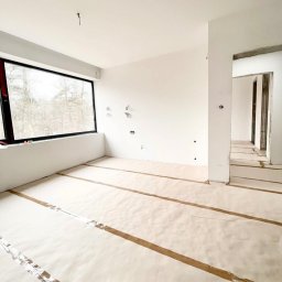 MD Construction - Wnętrze w stanie deweloperskim z dużym oknem PCV, zabezpieczona podłoga papierem i taśmą, widoczne instalacje elektryczne na ścianie.