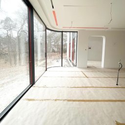 MD Construction - Wnętrze domu w trakcie wykańczania z zamontowanymi panoramicznymi oknami PCV z czarnymi ramami i czerwonymi akcentami, widok na zewnątrz na drzewa, podłoga zabezpieczona papierem malarskim.