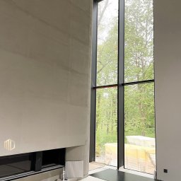 MD Construction - Wnętrze w trakcie wykańczania: surowa, jasna ściana z wbudowanym kominkiem i logo firmy, duże okno z czarną ramą z widokiem na zieleń, podłoga częściowo zabezpieczona folią.