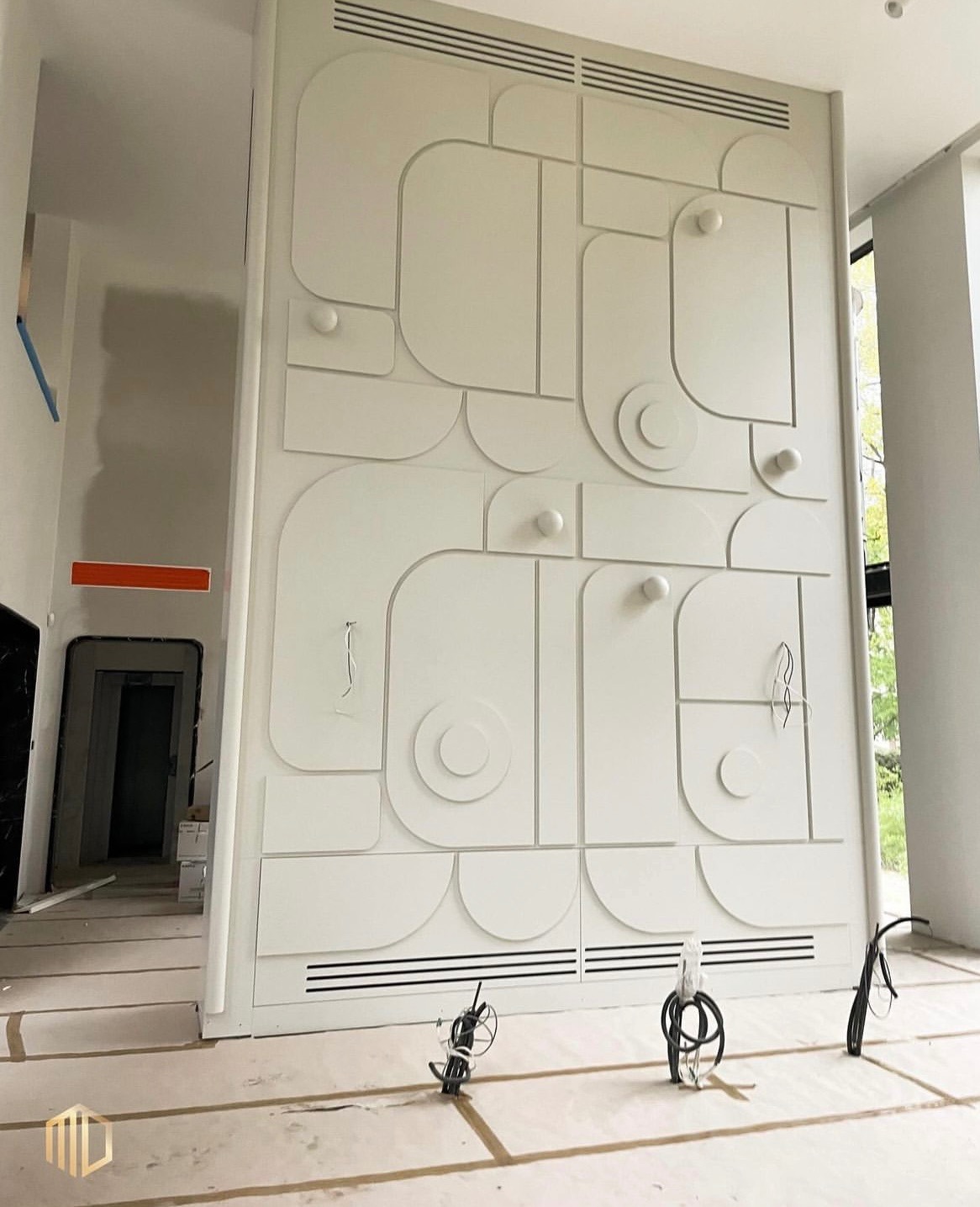 Wysoka, biała ściana z dekoracyjnym panelem 3D w geometryczne wzory, częściowo zamontowane oświetlenie punktowe, widoczne kable, podłoga zabezpieczona folią malarską, wnętrze w trakcie wykańczania.
