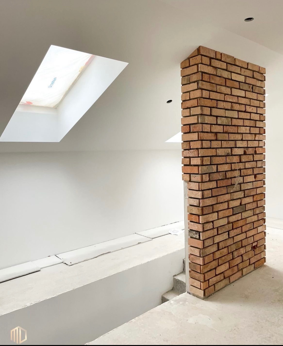 Wnętrze w trakcie wykończenia: ceglana kolumna na tle białych ścian, okna dachowe w skośnym suficie, surowy beton na podłodze i schodach.