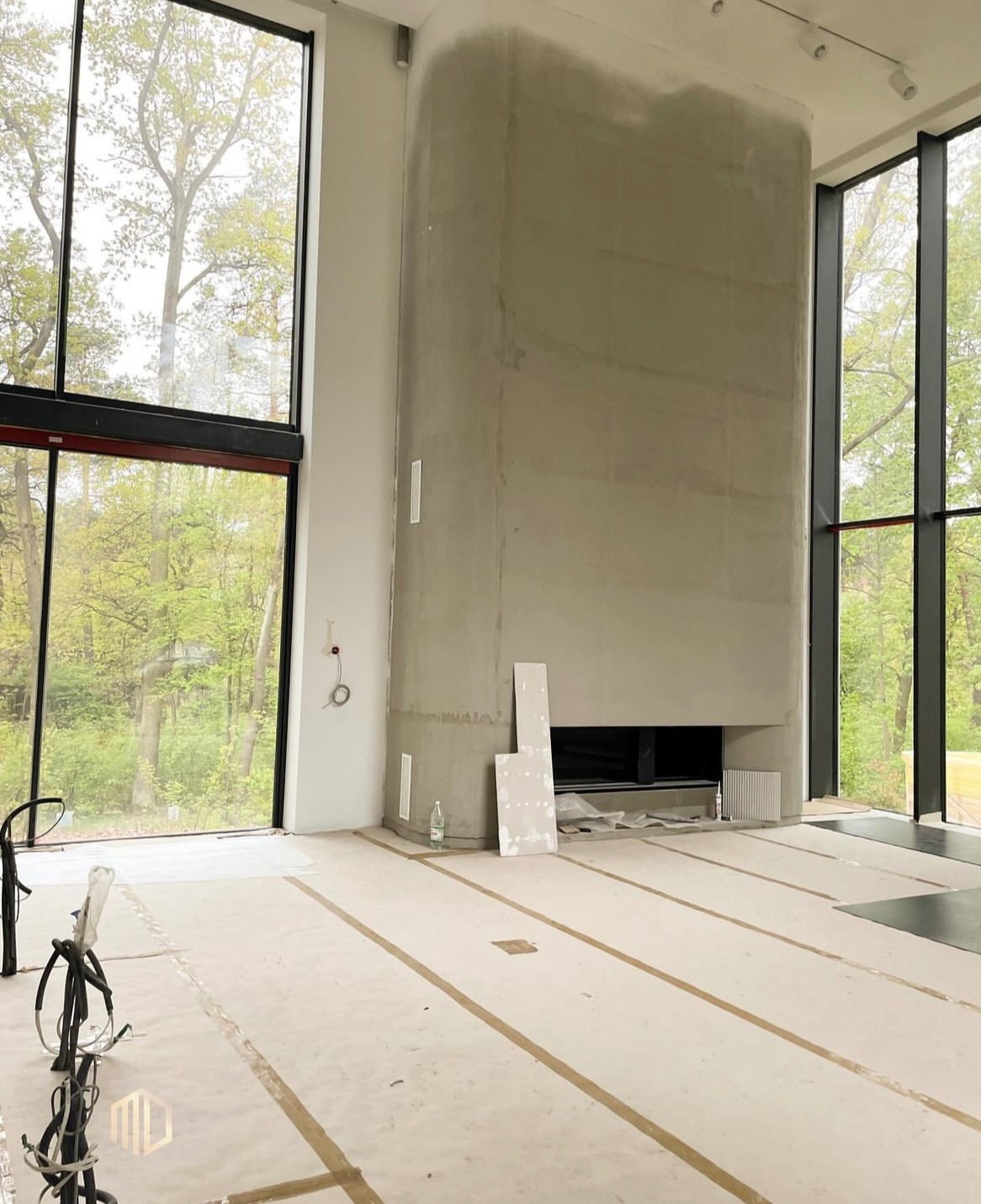 Wnętrze w trakcie wykończenia z dużymi oknami i surowym kominem z widocznymi śladami szpachlowania, zabezpieczona podłoga folią i taśmą, butelka wody i narzędzia budowlane w tle.
