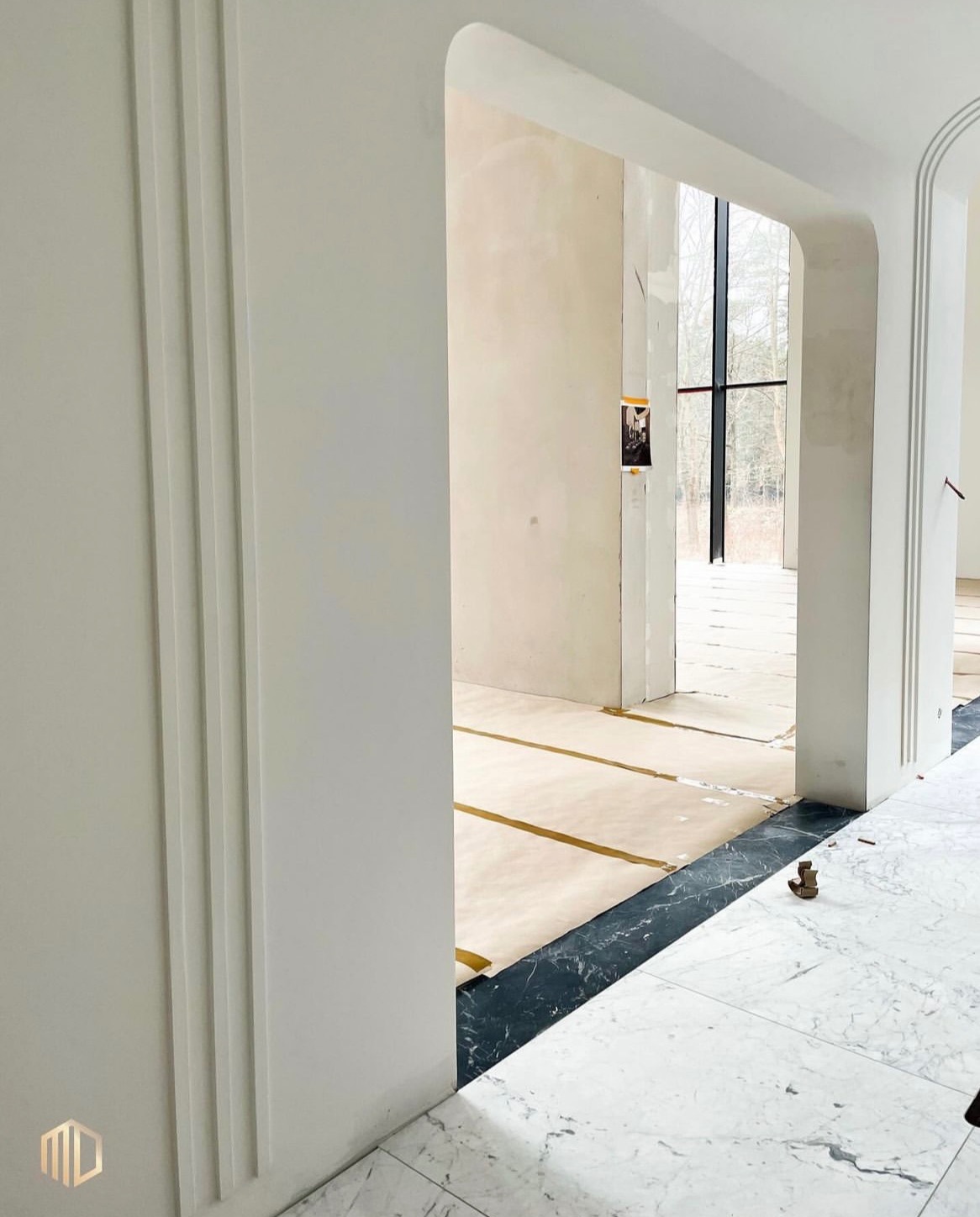 Wnętrze w trakcie wykańczania: białe ściany z dekoracyjnymi listwami, marmurowa podłoga z czarnym obramowaniem, widoczne zabezpieczenia podłogi, łukowe przejście do kolejnego pomieszczenia z dużym...