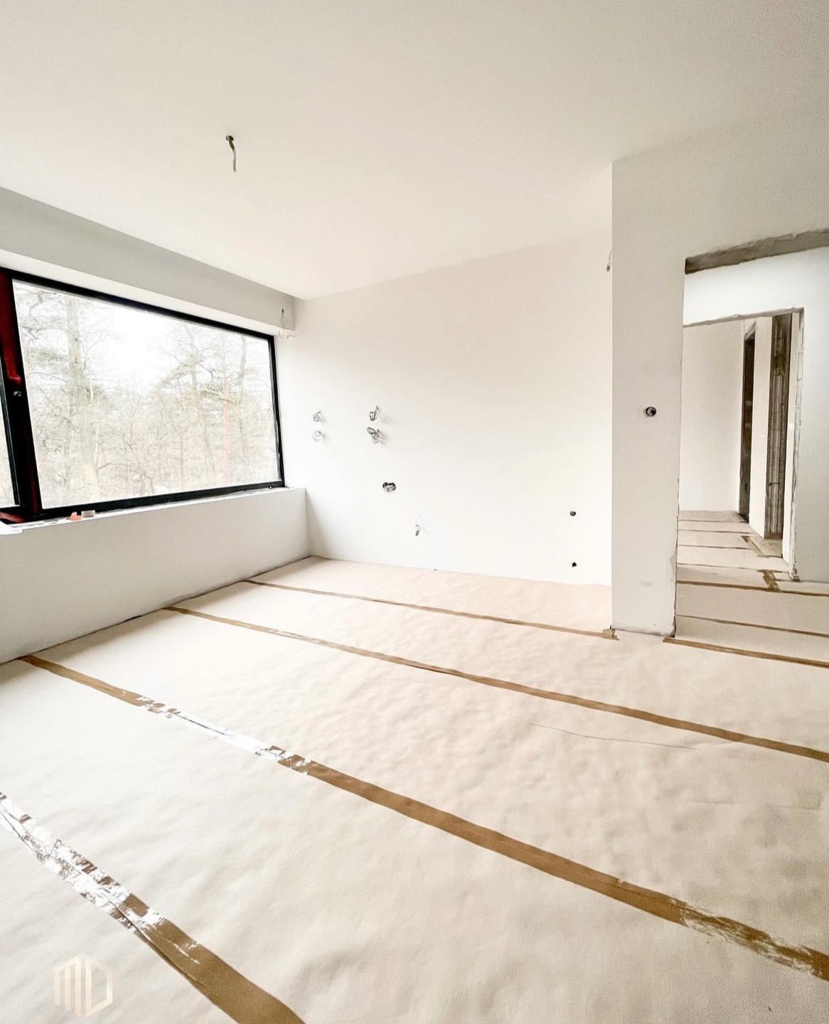 Wnętrze w stanie deweloperskim z dużym oknem PCV, zabezpieczona podłoga papierem i taśmą, widoczne instalacje elektryczne na ścianie.