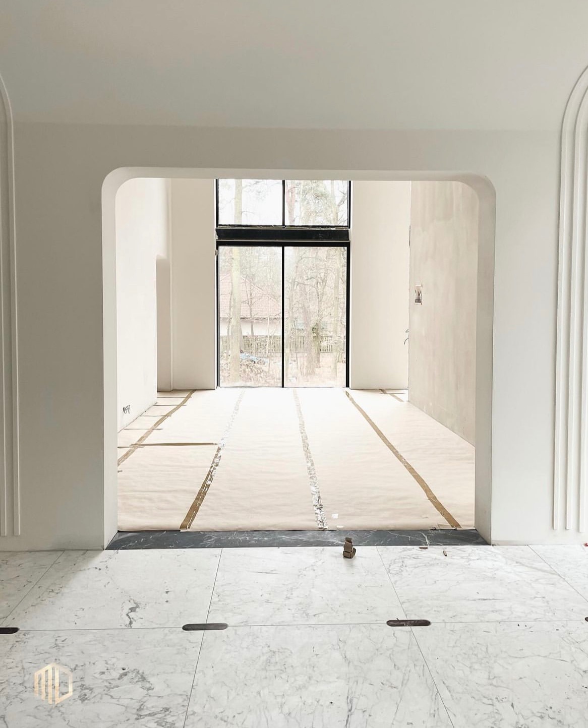 Wnętrze w trakcie wykańczania: marmurowa podłoga, zabezpieczona podłoga papierem malarskim, widok na okno z czarną ramą i drzewa w tle, białe ściany z dekoracyjnymi listwami.