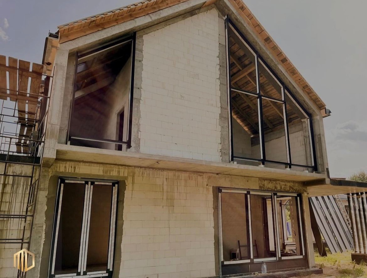 Surowy stan budynku z zamontowanymi oknami PCV z czarnymi ramami; widoczne rusztowanie i nieotynkowane ściany z betonu komórkowego.