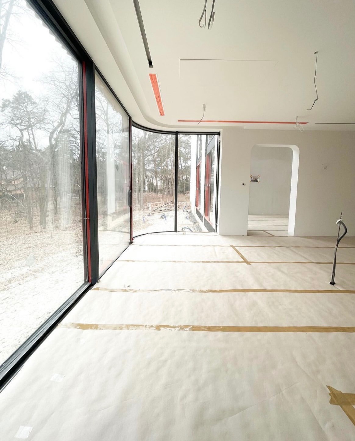 Wnętrze domu w trakcie wykańczania z zamontowanymi panoramicznymi oknami PCV z czarnymi ramami i czerwonymi akcentami, widok na zewnątrz na drzewa, podłoga zabezpieczona papierem malarskim.