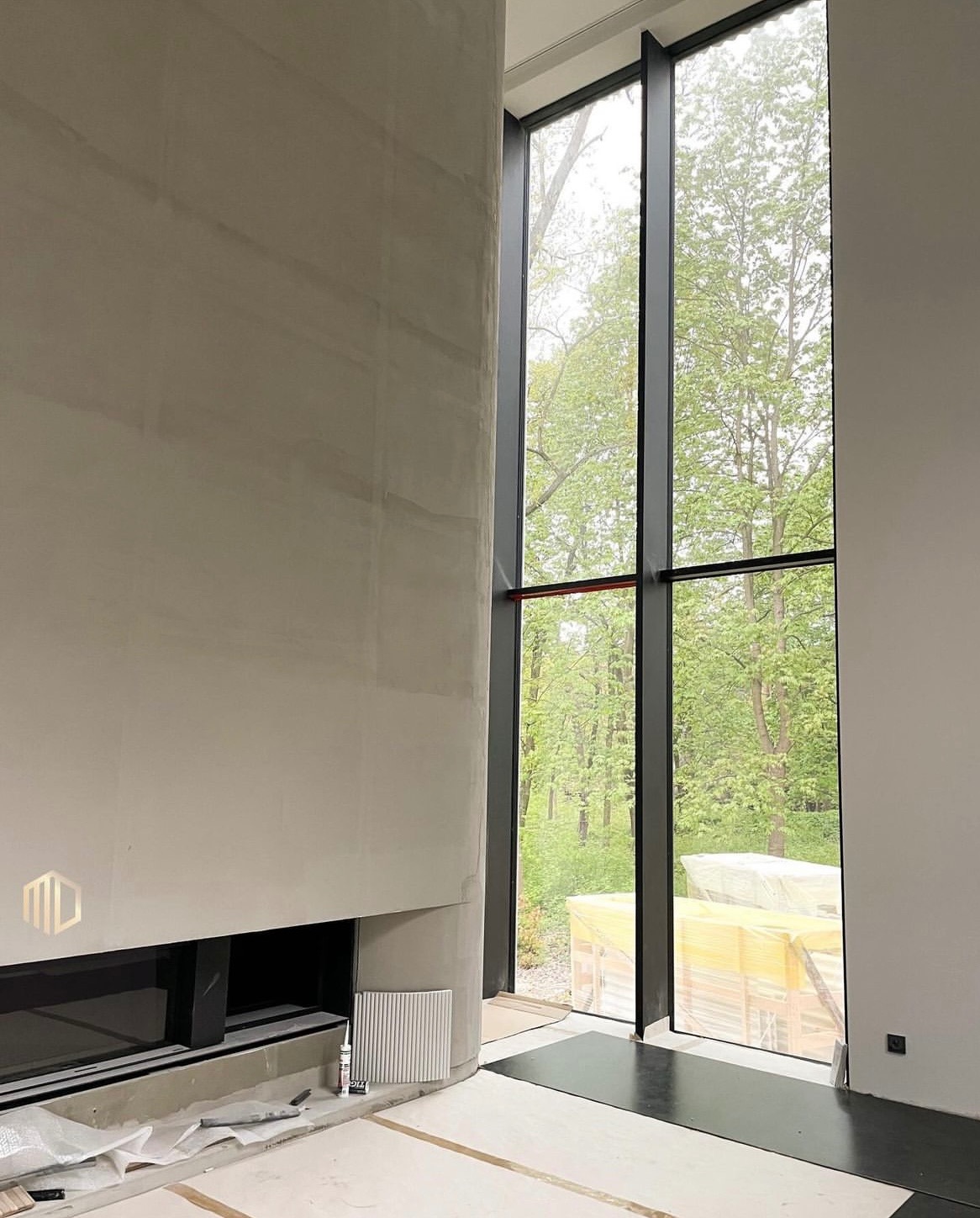 Wnętrze w trakcie wykańczania: surowa, jasna ściana z wbudowanym kominkiem i logo firmy, duże okno z czarną ramą z widokiem na zieleń, podłoga częściowo zabezpieczona folią.