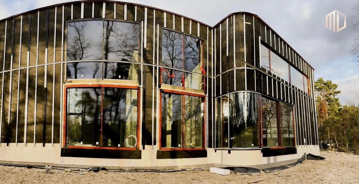 Budowa nowoczesnego domu w Warszawie: widoczne okna z czerwonymi ramami i elewacja w trakcie montażu, z widoczną izolacją i listwami.