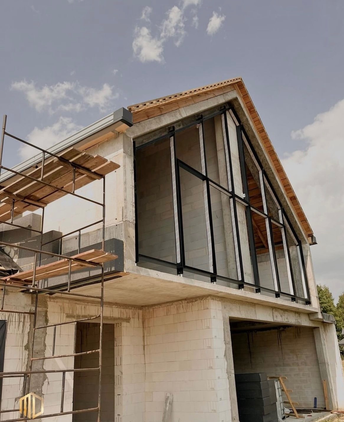 Budowa domu w stanie surowym otwartym: widoczne czarne ramy okienne w szczycie budynku, białe ściany z bloczków, rusztowania i drewniane elementy więźby dachowej pod szarym niebem.