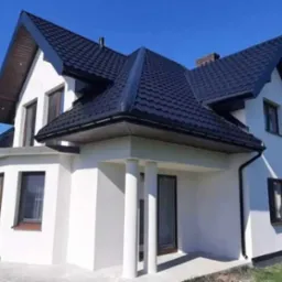 Nowoczesny dom jednorodzinny z białą elewacją i ciemnym dachem z blachodachówki, widok z poziomu gruntu w słoneczny dzień.