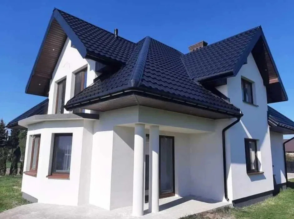 Nowoczesny dom jednorodzinny z białą elewacją i ciemnym dachem z blachodachówki, widok z poziomu gruntu w słoneczny dzień.