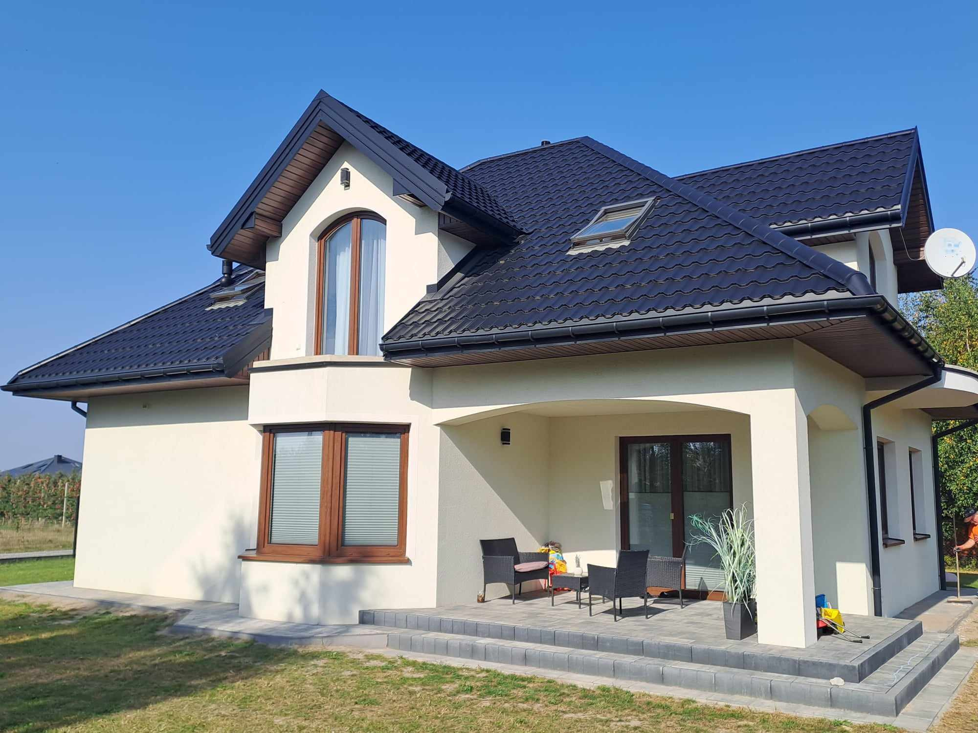 Jasny dom jednorodzinny z ciemną dachówką, widoczne okna z drewnianymi ramami i taras z meblami ogrodowymi, na trawniku zabawki dziecka.