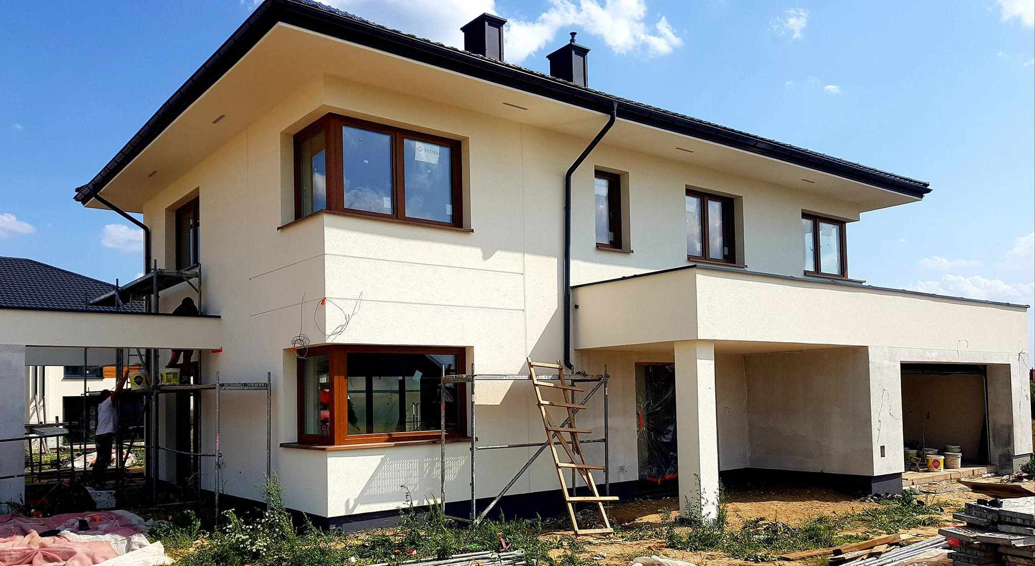 Dwukondygnacyjny dom jednorodzinny w trakcie prac elewacyjnych, widoczne rusztowania i drabina, jasna elewacja z ciemnymi oknami i dachem.