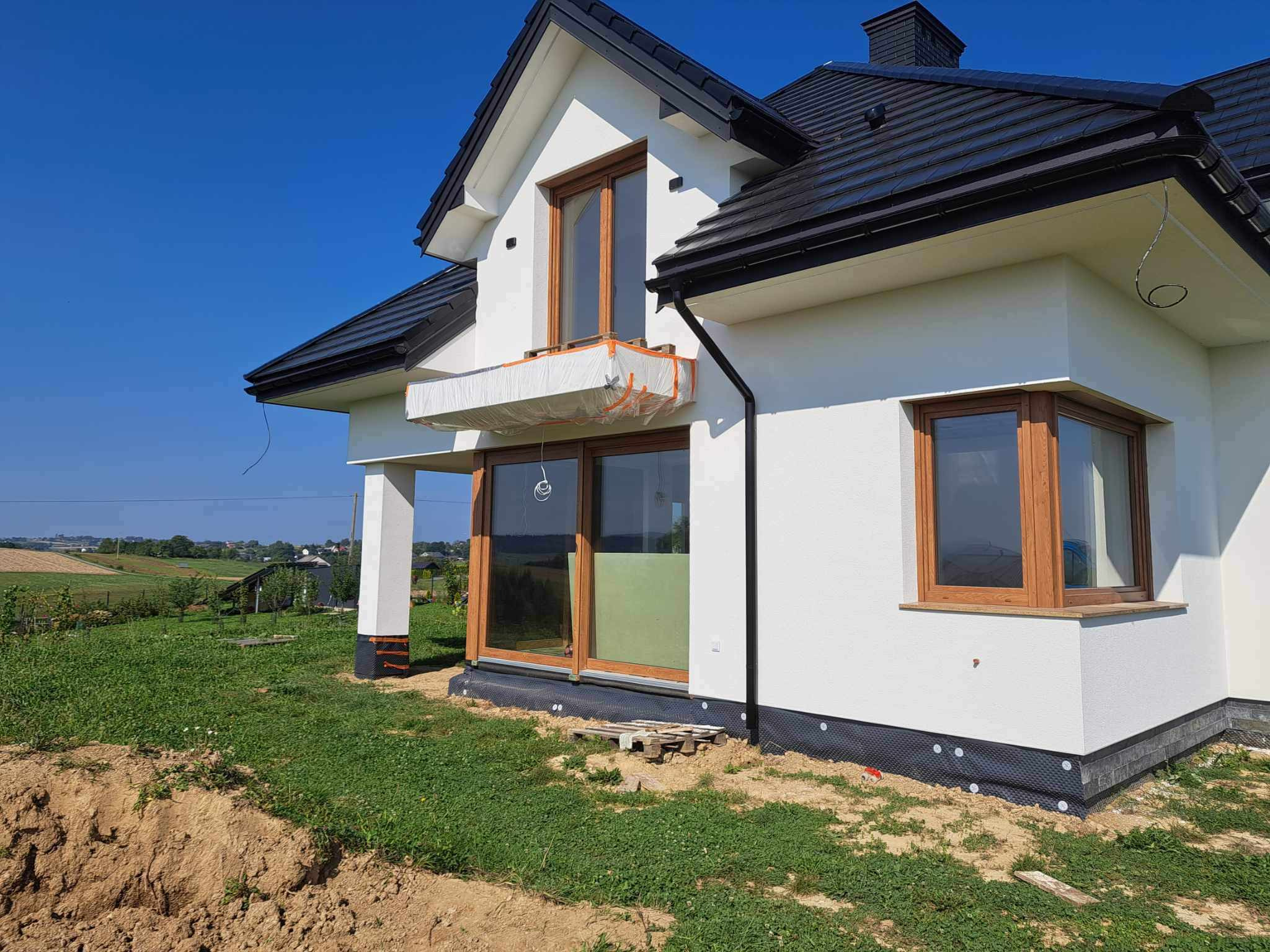 Dom jednorodzinny w trakcie prac elewacyjnych, widoczne okna z drewnianą ramą, ocieplony balkon zabezpieczony folią, czarna podbitka dachu i fragment krajobrazu w tle.