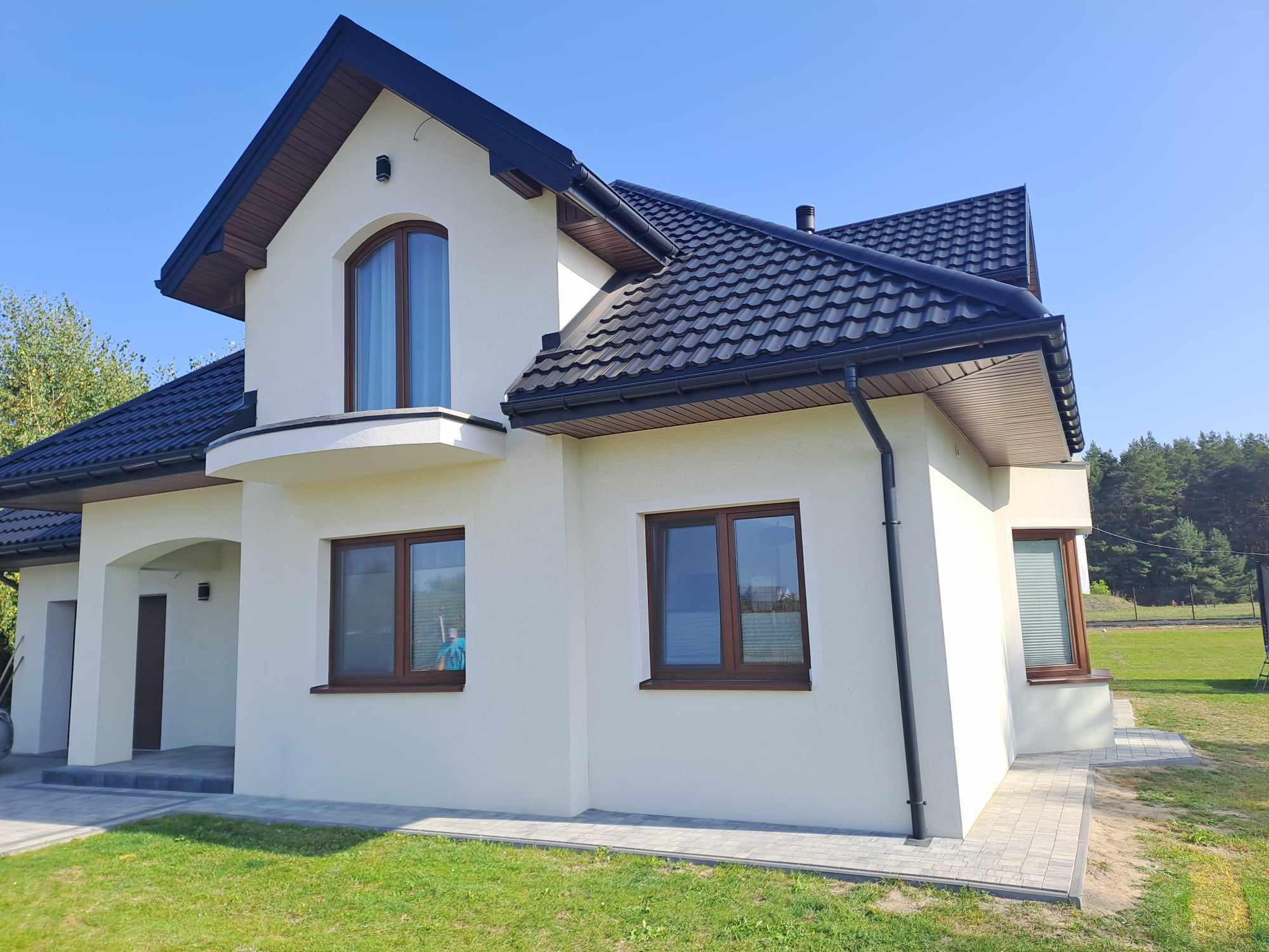 Nowoczesny dom jednorodzinny z jasną elewacją, ciemną dachówką i brązowymi oknami, sfotografowany w słoneczny dzień z perspektywy trawnika.
