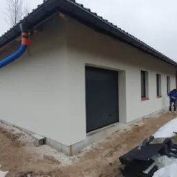 Świeżo otynkowana elewacja budynku z garażem i niebieską rurą spustową, inspektor w niebieskiej kurtce dokonuje inspekcji.